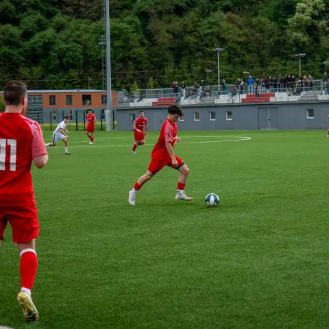 Spielszene: Ein Spieler in Rot dribbelt den Ball auf dem Kunstrasen, während ein Gegner in Weiß und Zuschauer auf der Tribüne zusehen.