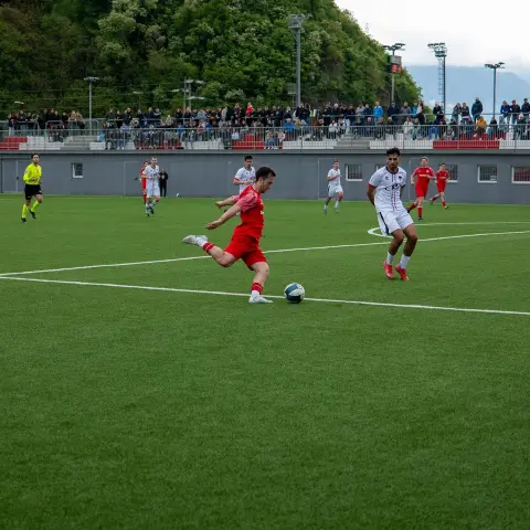 Spieler in roten Trikots schießt den Ball auf dem Kunstrasen, während ein Gegner in Weiß daneben läuft und Zuschauer auf der Tribüne zusehen.