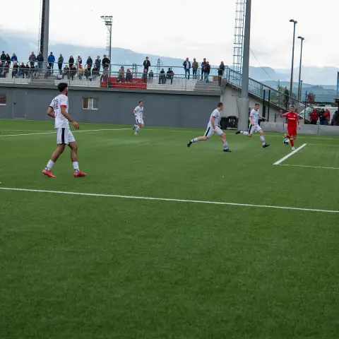 Spielszene auf dem Kunstrasen: Rote Mannschaft in roten Trikots gegen Weiß, ein Spieler führt den Ball, Zuschauer auf der Tribüne im Hintergrund.