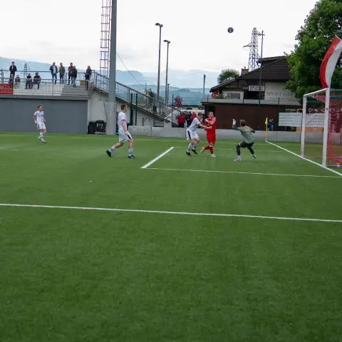 Spielszene auf dem Kunstrasen: Spieler in Rot und Weiß kämpfen vor dem Tor um den Ball, Zuschauer auf der Tribüne.