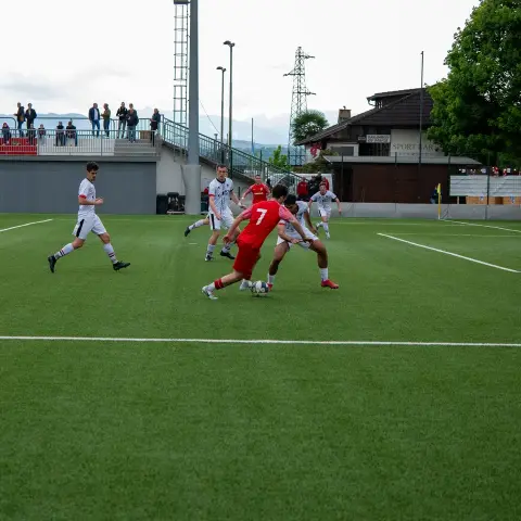 Spielszene auf dem Kunstrasenplatz: Spieler in Rot mit der Nummer 7 führt den Ball gegen zwei Gegenspieler in Weiß.