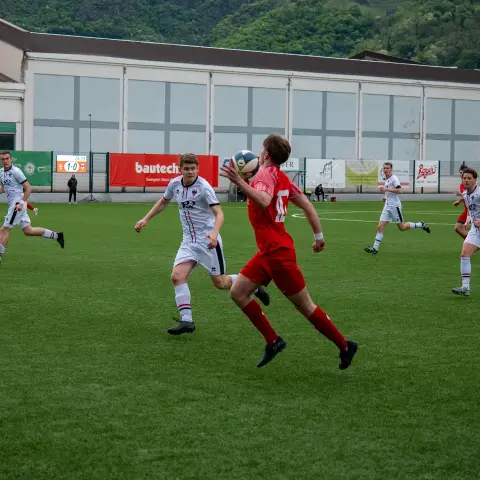 Ein Spieler in Rot jongliert den Ball im Sprung, während zwei Gegner in Weiß auf einem Kunstrasenplatz heranlaufen.