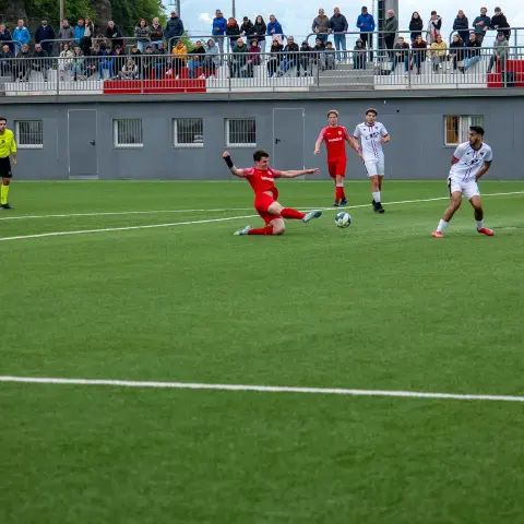 Spielszene: Ein roter Spieler grätscht am Ball, während weiß gekleidete Gegenspieler auf dem Kunstrasen zuschauen; Zuschauer auf der Tribüne im Hintergrund.