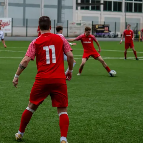 Spielszene: Ein Spieler in roter Trikot Nummer 11 steht mit dem Rücken zur Kamera, während ein Mitspieler in Rot den Ball am Boden führt, im Duell gegen Weiß.