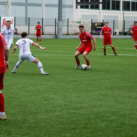 Spielszene: Ein roter Spieler führt den Ball gegen einen Gegner in weißem Trikot mit der Nummer 3, im Hintergrund weitere Spieler auf dem Kunstrasen.