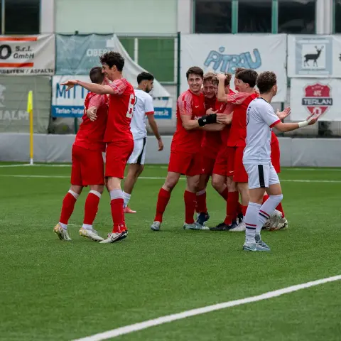 Rot gekleidete Spieler des AFC Terlan jubeln auf dem Kunstrasen dicht beieinander über ein Tor, im Hintergrund Bandenwerbung.