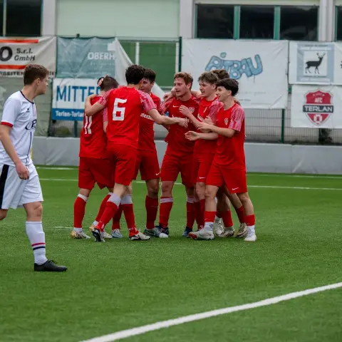 Spieler des AFC Terlan in roten Trikots jubeln eng umarmt nach einem Tor, im Hintergrund geht ein Gegenspieler in Weiß vorbei.