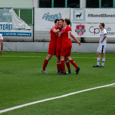 Drei Spieler des AFC Terlan in roten Trikots jubeln eng zusammen auf dem Kunstrasenplatz, ein Gegenspieler in Weiß steht im Hintergrund.