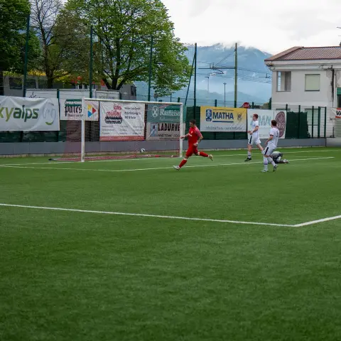 Spielszene auf dem Kunstrasen: Ein Spieler in Rot läuft auf das Tor zu, während Gegner in Weiß zusehen; Banden mit Sponsorenwerbung im Hintergrund.