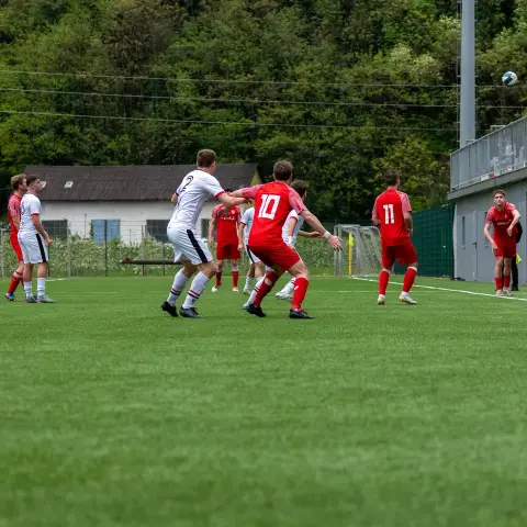 Spielszene auf einem Kunstrasenplatz: Ein Spieler in Rot mit Nummer 10 steigt zum Kopfball hoch, daneben ein Gegner in Weiß, im Hintergrund Zuschauer und Waldhang.