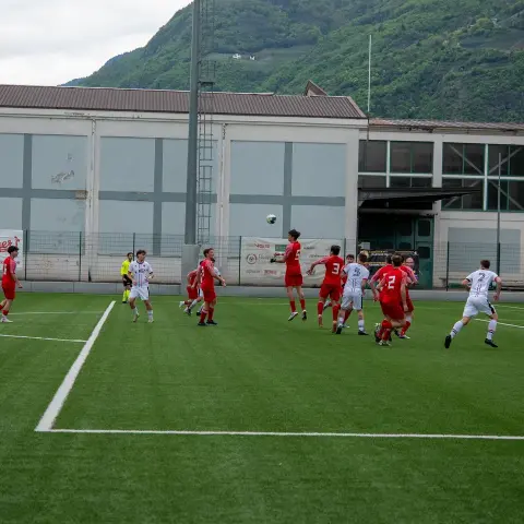 Spielszene auf dem Kunstrasen: Spieler in Rot und Weiß kämpfen im Strafraum um einen hohen Ball, im Hintergrund Berghang und Sponsorenbanden.