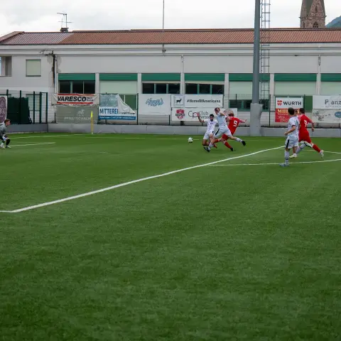 Spielszene auf dem Kunstrasen: Spieler in Rot und Weiß kämpfen um den Ball, im Hintergrund Bandenwerbung und ein Vereinsgebäude.