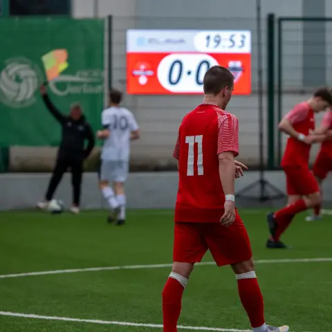 Spieler in roter Trikotnummer 11 steht auf dem Kunstrasen, im Hintergrund Spieler in Rot und Weiß und ein Anzeigetafel-Stand 0:0 um 19:53.
