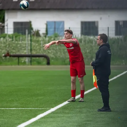 Spieler in roter Trikot wirft den Ball vom Seitenaus ein, daneben steht ein Linienrichter mit Fahne auf dem Kunstrasen.