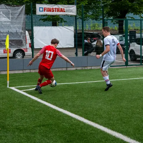 Spielszene am Eckfahnen: Ein Spieler in Rot mit Nummer 10 führt den Ball, ein Gegenspieler in Weiß mit Nummer 5 läuft ihm nach.