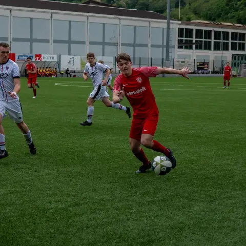 Spielszene: Ein Spieler in roter Trikot mit Sponsorenaufdruck führt den Ball, während zwei Gegner in Weiß auf dem Kunstrasen nachsetzen.