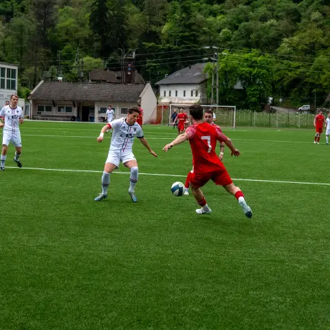 Spielszene: Ein Spieler in Rot mit der Nummer 7 führt den Ball gegen einen Gegenspieler in Weiß auf dem Kunstrasen.