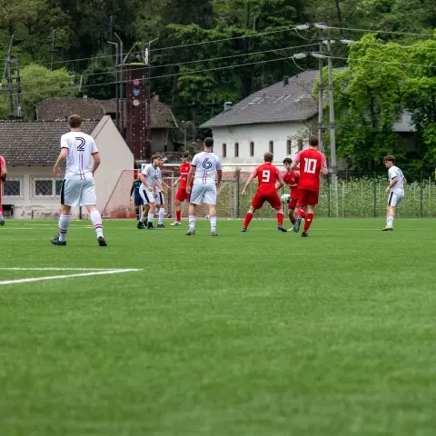 Spielszene auf dem Kunstrasen: Rot gekleidete Spieler stehen in Ballnähe gegen weiß gekleidete Gegner, im Hintergrund Bäume und Häuser.