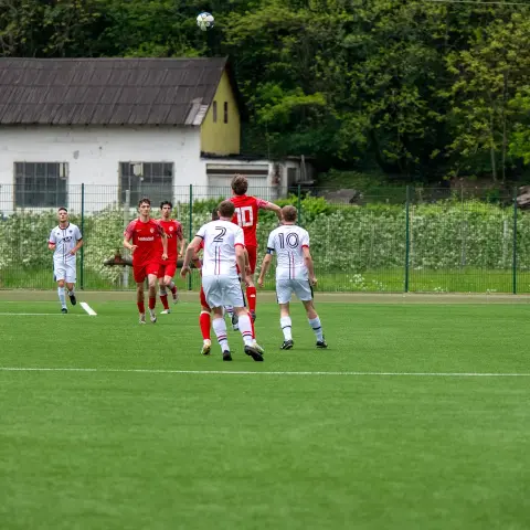 Spielszene auf einem Kunstrasenplatz: Ein Spieler in Rot mit Nummer 10 springt zum Kopfball, umringt von Spielern in Weiß und Rot, im Hintergrund ein Haus und Bäume.