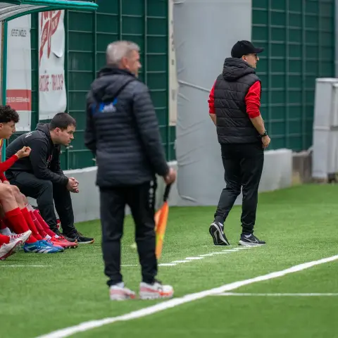 Trainer und Ersatzspieler in roten Trikots am Spielfeldrand beobachten ein Spiel auf Kunstrasen, im Vordergrund ein Trainer mit Fahne.