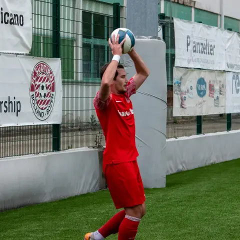 Spieler in roter Trikot wirft den Ball zum Einwurf an der Seitenlinie, im Hintergrund Werbebanner von Sponsoren auf dem Kunstrasenplatz.