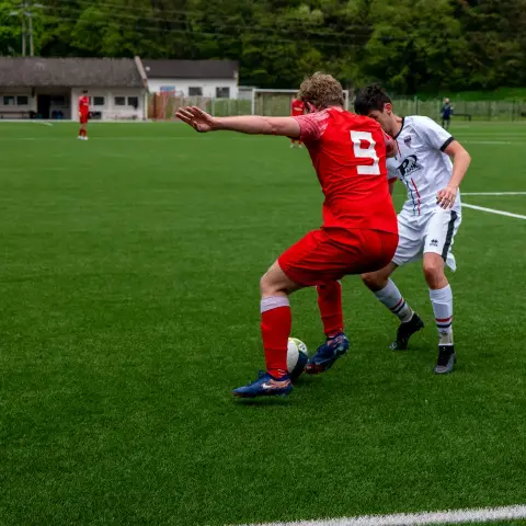 Spielszene: Ein Spieler in roter Trikotnummer 9 schirmt den Ball gegen einen Gegner in weißem Trikot mit Sponsorlogo ab, im Hintergrund weitere Spieler auf dem Kunstrasen.