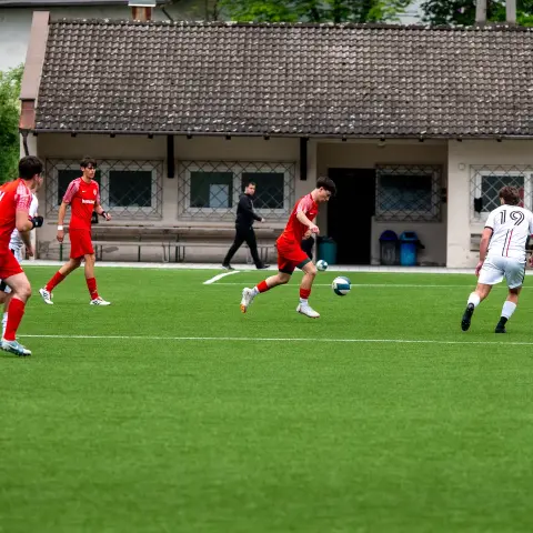 Spielszene auf dem Kunstrasen: Ein Spieler in Rot führt den Ball, während ein Gegenspieler in Weiß mit der Nummer 19 und der Schiedsrichter in Gelb zusehen.