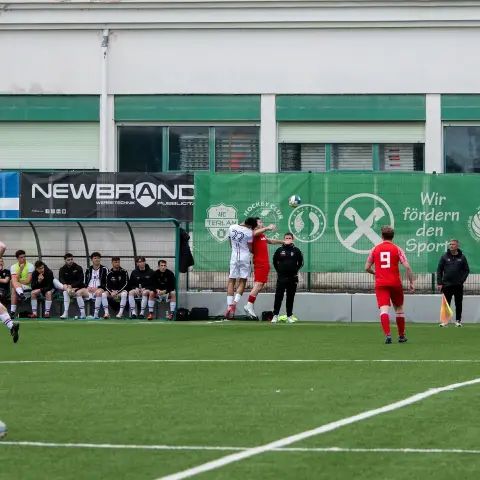 Zwei Spieler in Weiß und Rot springen beim Kopfball um den Ball, davor Sponsorenbanden und Bank am Spielfeldrand des AFC Terlan.