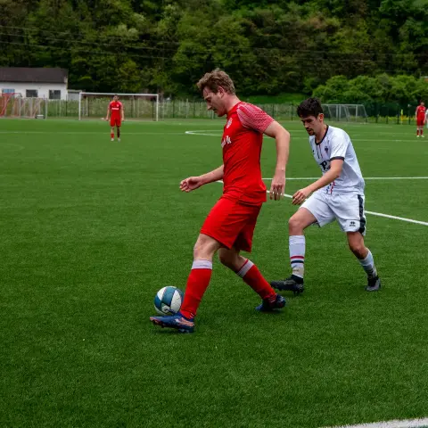 Spielszene: Ein Spieler in roter Trikot führt den Ball am Fuß, während ein Gegenspieler in Weiß versucht, ihn zu stören, auf einem grünen Kunstrasenplatz.