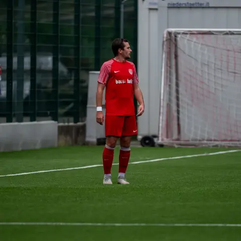 Ein Spieler in roter Vereinskleidung steht auf dem Kunstrasen neben dem Tor und blickt zur Seite, im Hintergrund ein Sponsor-Schriftzug auf der Bande.