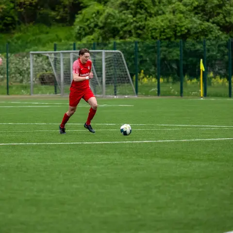 Spielszene auf dem Kunstrasen: Ein Spieler in roten Trikot führt den Ball, ein Gegenspieler in Weiß läuft von links heran.