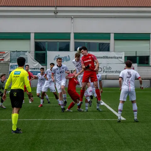 Kopfballduell im Strafraum: Ein Spieler in Rot springt hoch zum Ball, umringt von Spielern in Weiß, Torwart und Schiedsrichter in Gelb verfolgen die Szene.
