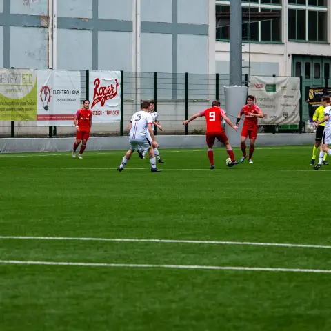 Spielszene auf dem Kunstrasen: Spieler in Rot und Weiß kämpfen um den Ball, im Hintergrund Werbebanden und Zuschauerbank.