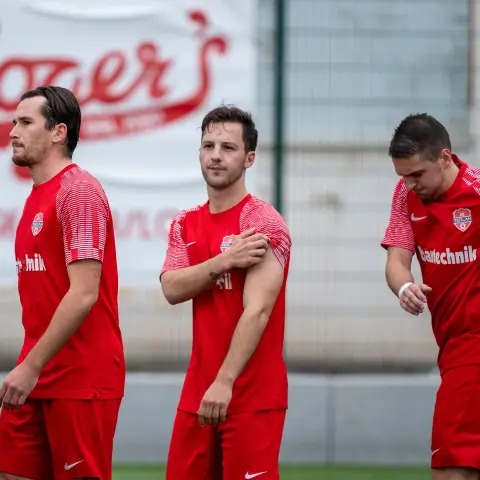 Rote Spieler des AFC Terlan stehen am Spielfeldrand vor einer Bbande mit Rozzer-Werbung, im Vordergrund ein Spieler mit Blick zur Seite.