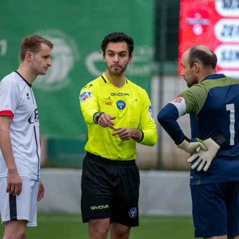 Schiedsrichter in neongelber Trikot zeigt im Gespräch mit einem Torwart in blau-grüner Trikot auf der Spielfläche, daneben zwei Spieler in Weiß und Grau.