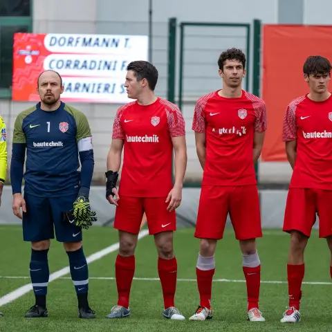 Spieler des AFC Terlan in roten Trikots mit Bautechnik-Sponsor stehen vor einem Spiel Seite an Seite, links Schiedsrichter in Neongelb und Torhüter in Dunkelblau.