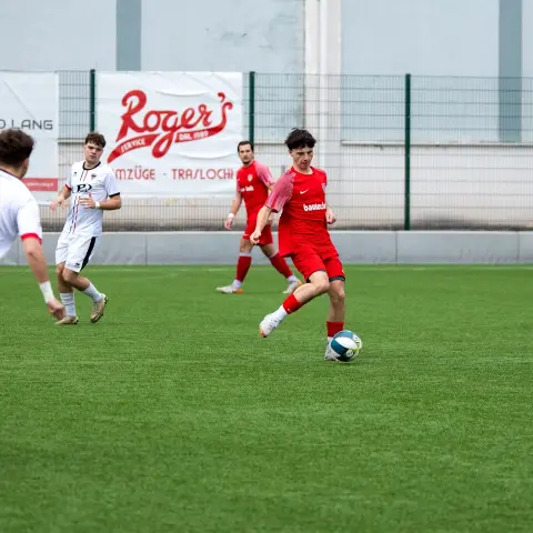 Spielszene: Ein Spieler in rotem Trikot führt den Ball am Rand eines Kunstrasenplatzes, im Vordergrund ein Gegenspieler in Weiß mit der Nummer 3.