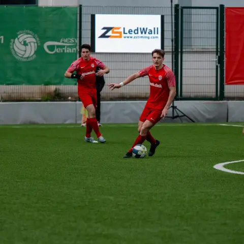 Ein Spieler in roter Trikot führt den Ball auf dem Kunstrasen, während ein Gegenspieler in Weiß von rechts heranläuft.