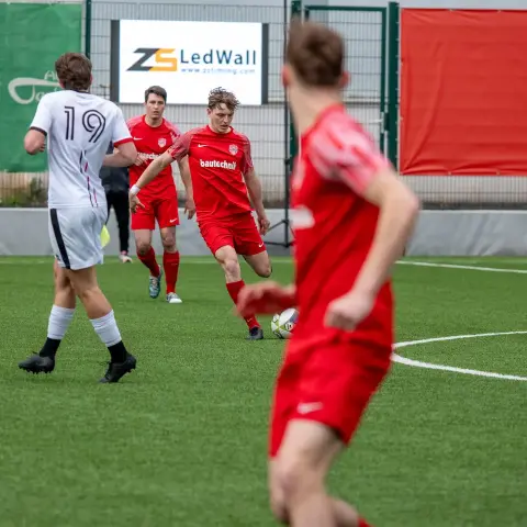 Spielszene auf dem Kunstrasen: Ein Spieler in Rot führt den Ball, im Vordergrund ein Mitspieler in Rot, links ein Gegner in Weiß mit der Nummer 19.
