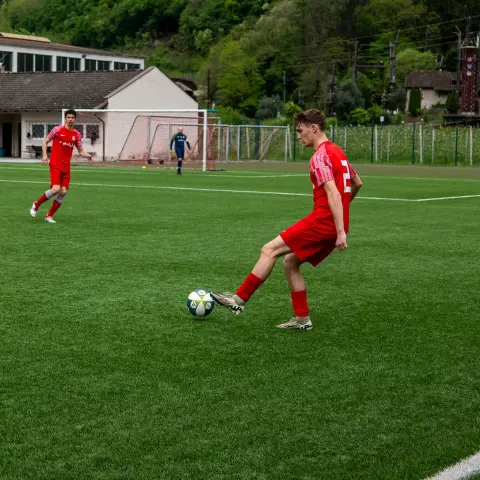 Ein Spieler in roter Trikot und roter Hose schießt auf einem grünen Kunstrasenplatz den Ball, daneben laufen Mitspieler und ein Torwart im Hintergrund.