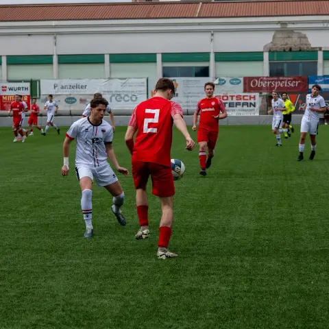 Spielszene: Spieler in rotem Trikot mit der Nummer 2 führt den Ball, ein Gegenspieler in Weiß attackiert auf dem Kunstrasen.