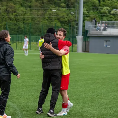 Zwei Spieler umarmen sich nach dem Spiel auf dem Kunstrasen, im Hintergrund geht eine Frau in schwarzer Jacke vorbei.