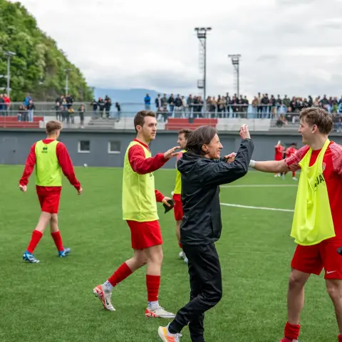 Spieler in roten Trikots und gelben Leibchen klatschen auf dem Kunstrasenplatz mit einer Trainerin in schwarzer Jacke ab, im Hintergrund Zuschauer auf der Tribüne.