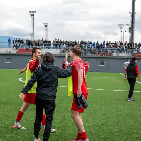 Rote Spieler klatschen nach dem Spiel auf dem Kunstrasen mit einem Betreuer in schwarzer Jacke ab, im Hintergrund sitzen Zuschauer auf der Tribüne.