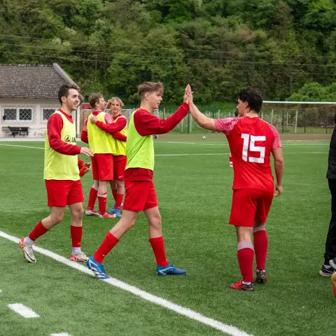 Spieler in roten Trikots klatschen sich auf dem Rasen ab, daneben wartet eine Gruppe in roten Trikots mit gelben Leibchen am Spielfeldrand.