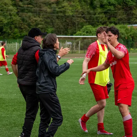 Zwei Betreuer in dunkler Kleidung klatschen am Spielfeldrand mit Spielern des AFC Terlan in roten Trikots ab, im Hintergrund trainieren weitere Spieler.