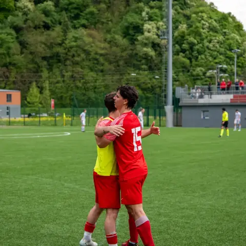 Zwei Spieler des AFC Terlan in roten Trikots umarmen sich nach einer Szene auf dem Rasen, im Hintergrund Zuschauer auf der Tribüne.