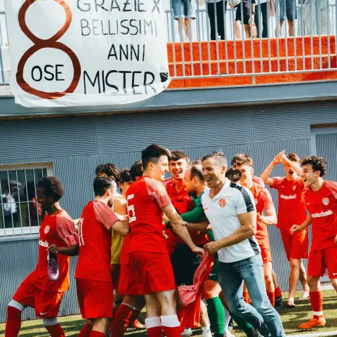 Rote Spieler des AFC Terlan jubeln und umarmen einen Trainer in weißem Poloshirt vor einem Banner mit 'Grazie x bellissimi anni, 8 ose mister'.