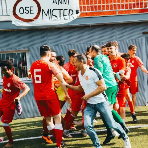 Terlaner Spieler in roten Trikots jubeln am Spielfeldrand, im Vordergrund ein Trainer in Weiß, daneben ein Banner mit der Aufschrift 'Grazie x bellissimi anni Mister'.