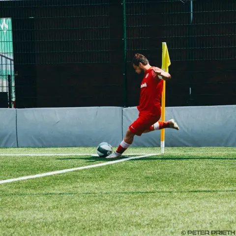 Ein Spieler in roter Trikot führt an der Eckfahne einen Eckstoß aus, der Ball liegt auf dem Rasen vor ihm.